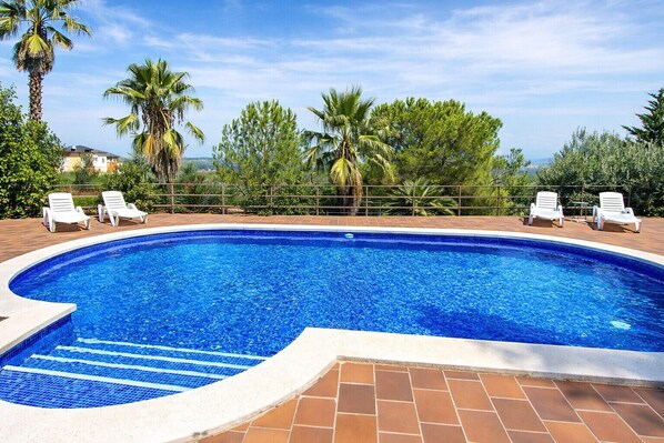 Una piscina con borde de azulejos, rodeada por un patio de ladrillo. Hay sillas reclinables blancas a lo largo de la piscina.