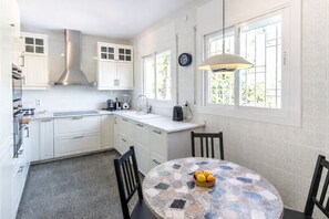 A modern kitchen with white cabinetry, a stainless steel range hood, a built-in oven, and a coffee machine. There is a round table with a mosaic tablecloth and a bowl of fruit.