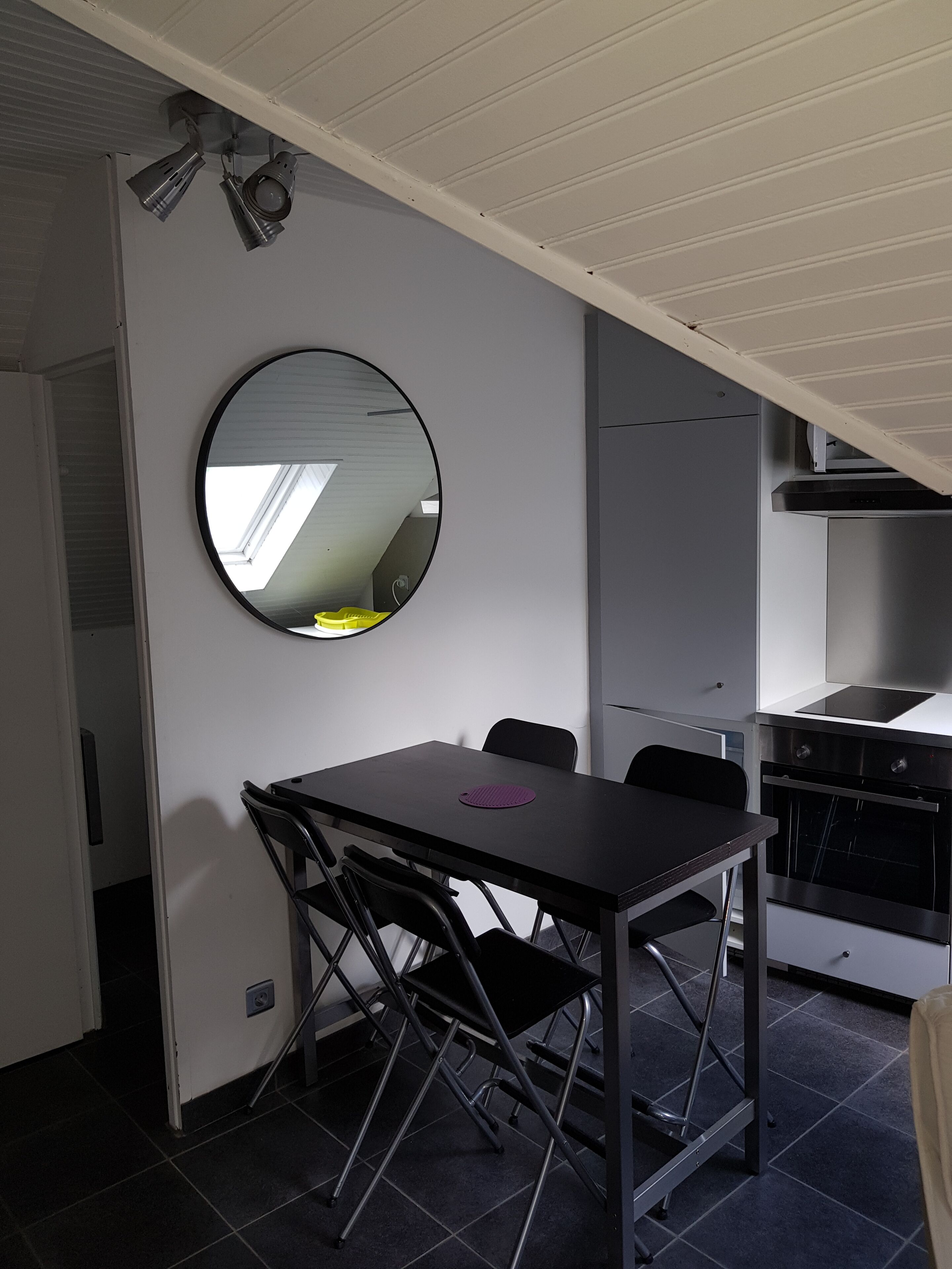 A compact kitchen with a dining area, including a foldable table and chairs, a round mirror, and a built-in oven.