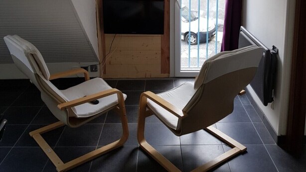 A small, sloped-roofed room with two wooden-framed chairs, a flat-screen TV mounted on the wall, and a window with a view of parked cars outside. - St Lary-Vignec Nice little apartment ideally located (St Lary- Vignec)