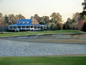 A large house with a blue roof and multiple chimneys, situated next to a body of water and a golf course.