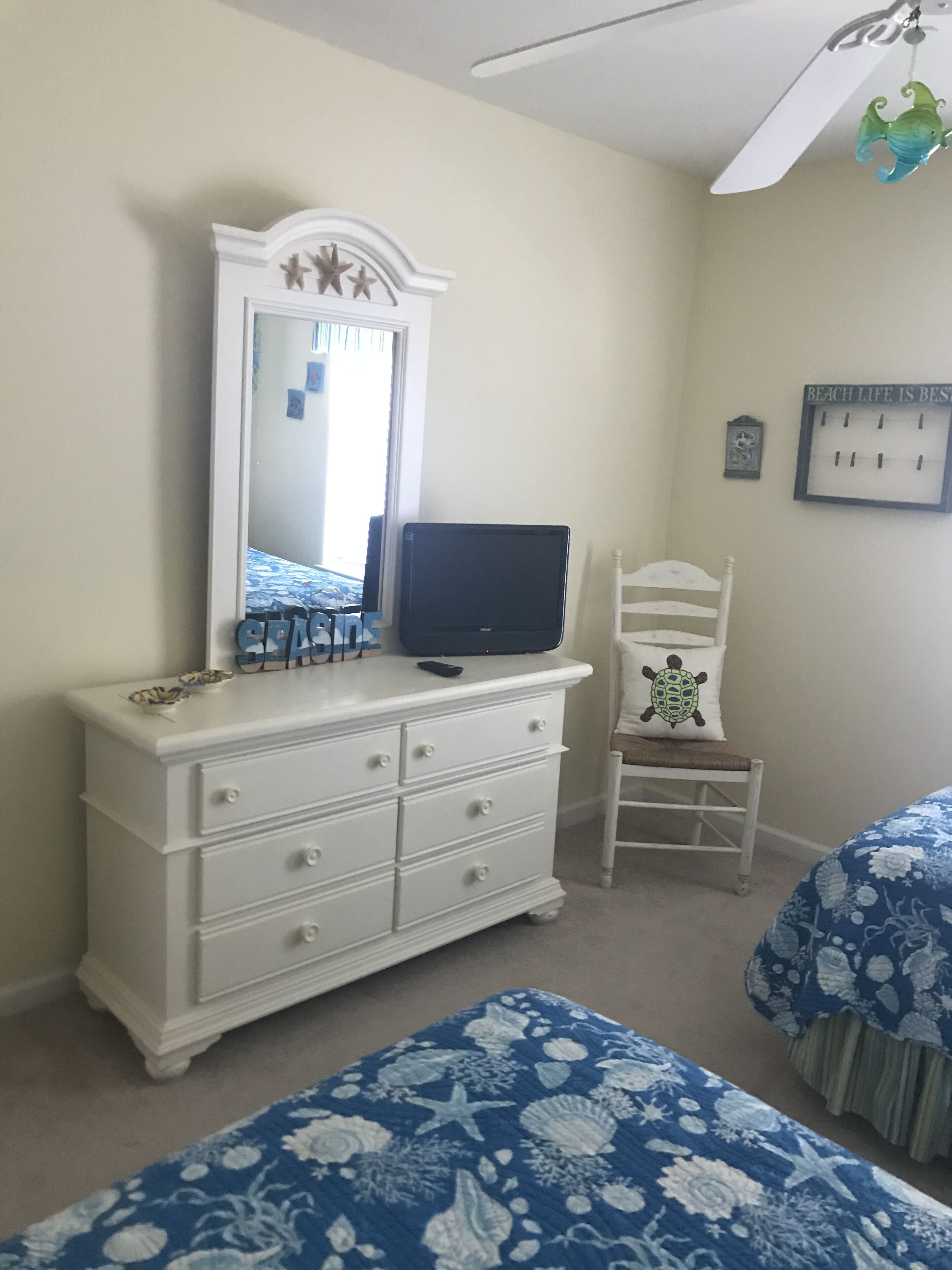 Une chambre avec une commode blanche, un téléviseur posé dessus, une chaise ornée d’un coussin à motif de tortue et un lit avec une literie à fleurs bleues.