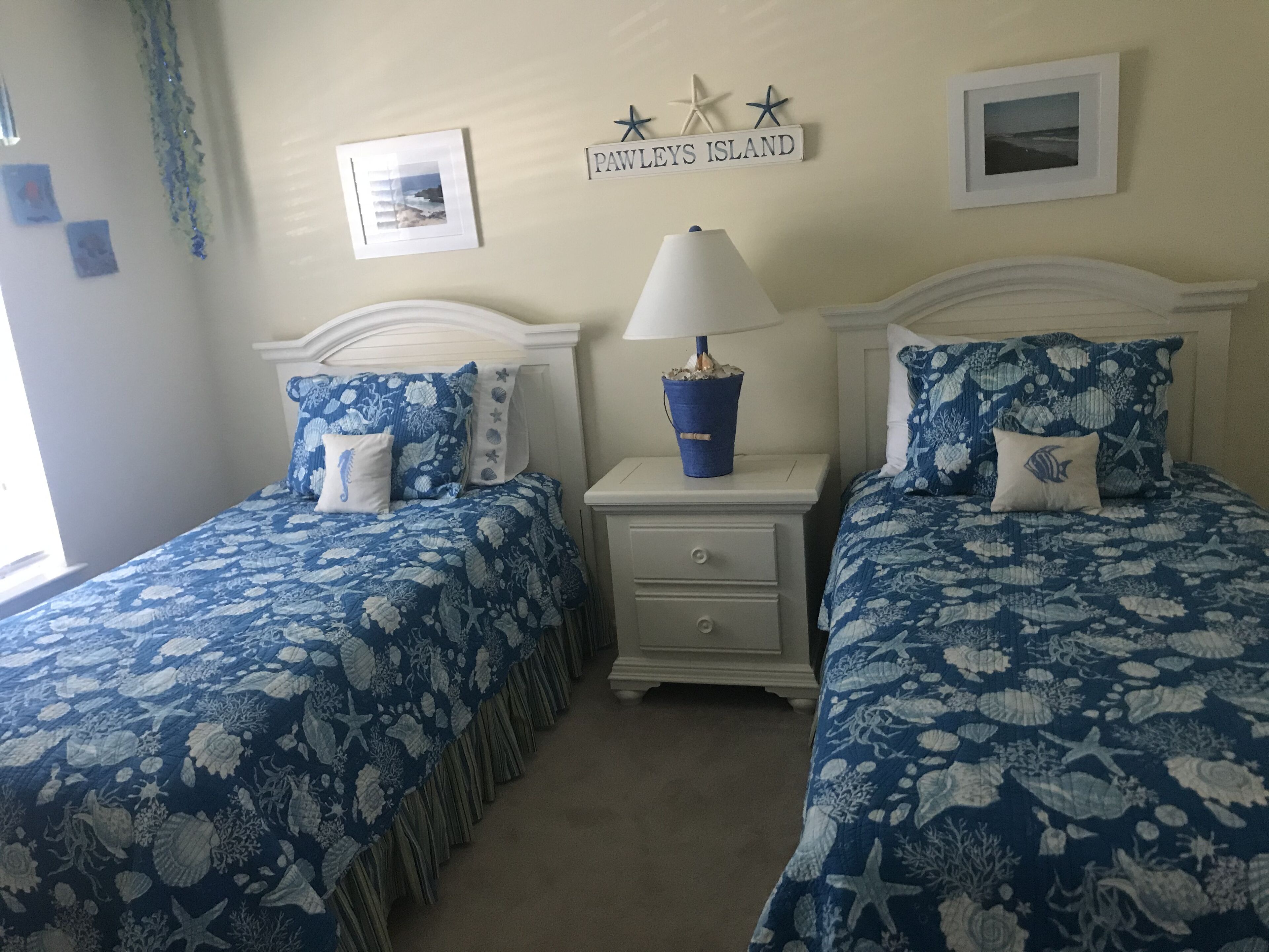 Ein Schlafzimmer mit zwei Betten, jeweils mit einer blauen, floralen Bettdecke und passenden Kissen. Zwischen den Betten befindet sich ein Nachttisch mit einer Lampe und einem kleinen Dekorationsgegenstand. An der Wand hängt ein Schild mit der Aufschrift „PAWLEYS ISLAND“ und ein gerahmtes Bild.