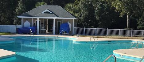 A clear swimming pool with a covered pavilion and lounge chairs in the background.