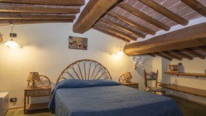 Une chambre avec un plafond en bois, un lit recouvert d’une courtepointe bleue, deux tables de chevet avec des lampes, une chaise et une étagère avec un cadre photo.