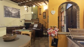 Un coin repas avec un piano, des chaises en bois, une table avec un panier, et un mur orné d’un tableau encadré et d’une décoration en forme de cœur suspendue.