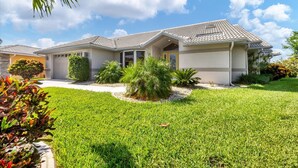 A two-story house with a tiled roof, a garage, and a well-maintained lawn with various shrubs and a palm tree. - Punta Gorda Isles 3 Bedroom Private Pool+Spa Home on Canal (Punta Gorde)