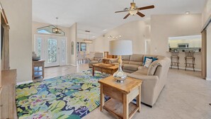 A spacious living room with a beige sofa, a wooden coffee table, and a colorful rug. There is a ceiling fan, a lamp, and a kitchen area visible in the background. - Punta Gorda Isles 3 Bedroom Private Pool+Spa Home on Canal (Punta Gorde)
