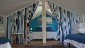 Un dormitorio con techo inclinado, cama con ropa de cama azul, cortinas a rayas y una mesita con una lámpara.