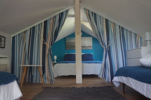 Un dormitorio con techo inclinado, cama con ropa de cama azul, cortinas a rayas y una mesita con una lámpara.
