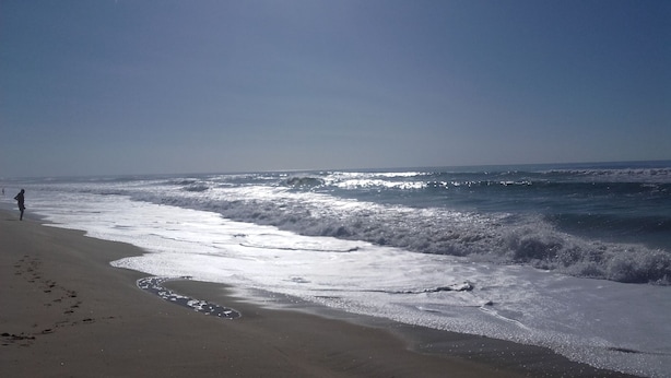 Una playa con olas acercándose a la orilla, un cielo despejado y una persona caminando por la playa.