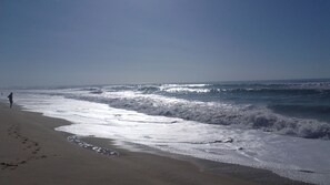 Una playa con olas acercándose a la orilla, un cielo despejado y una persona caminando por la playa.
