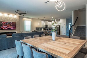 A modern living room with a large wooden dining table, grey upholstered sofas, and a flat-screen TV mounted on a textured wall.