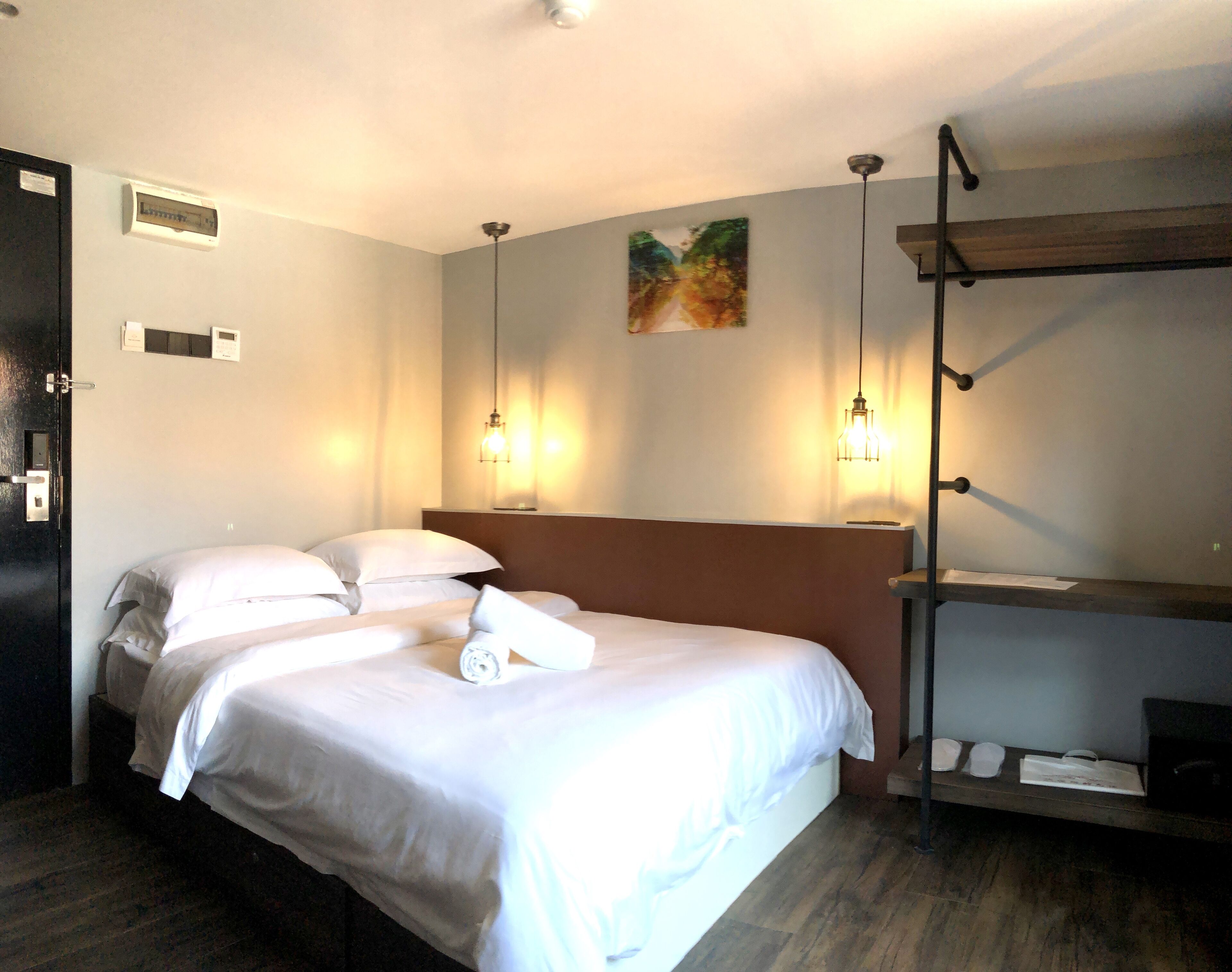 Habitación de hotel con cama, mesitas de noche, cabecero, un cuadro en la pared y un estante con un espejo y una bolsa.