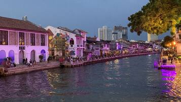 A riverside walkway with buildings on either side, a bridge, and a tree.