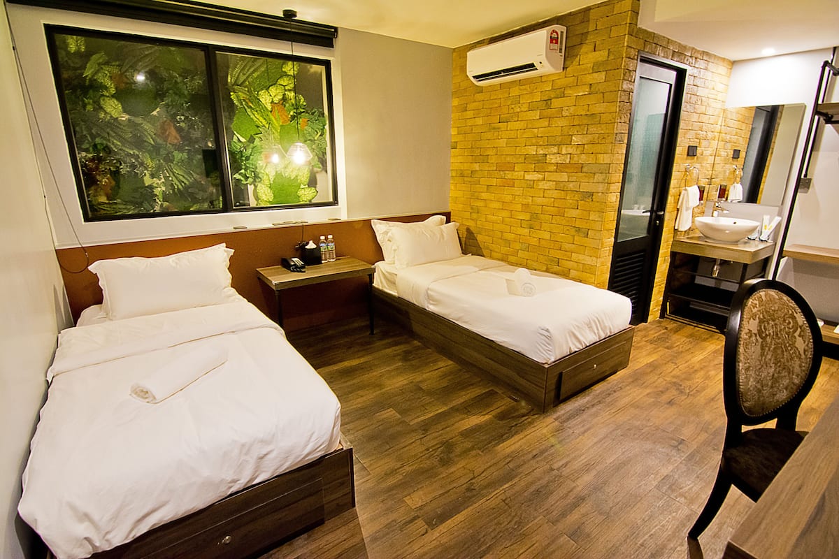 Een hotelkamer met twee bedden, een houten vloer, een accentmuur van baksteen, een raam met planten en een badkamer met een wastafel en spiegel.