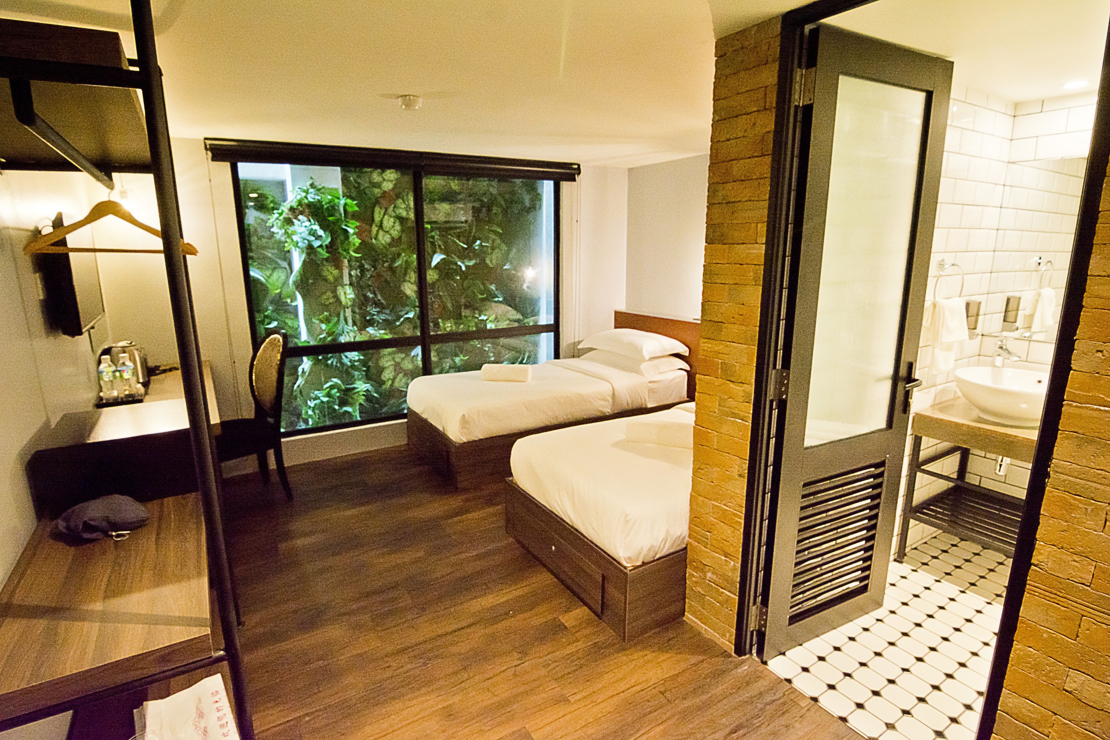 Habitación de hotel con dos camas, piso de madera, un ventanal con vistas a zonas verdes, baño con lavamanos y espejo, y un escritorio de madera con una silla.