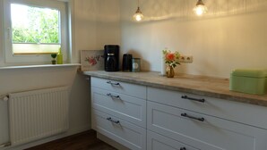 A kitchen with white cabinets, a window with blinds, a radiator, a coffee maker, a vase with flowers, and pendant lights. - VALUE HOF | cozy holiday house (Bracht)
