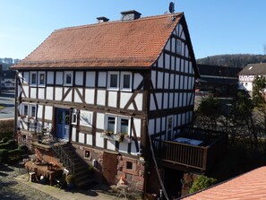 Une maison traditionnelle à colombages, dotée d’un toit à forte pente, d’un escalier extérieur menant à l’entrée et d’un petit espace extérieur aménagé avec une table et des chaises.