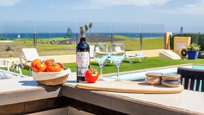 Et bord med vinflaske, glas, tomater og syltetøjsglas på en terrasse med udsigt over en pool og havet.