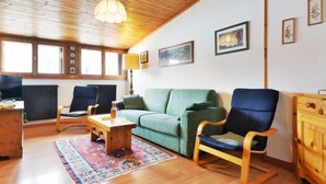 Ein gemütliches Wohnzimmer mit Holzboden, einem grünen Sofa, einem blauen Sessel, einem Holztisch und einem bunten Teppich.