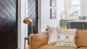 A living room with a brown leather sofa, a patterned throw pillow, a floor lamp, and a coffee table with books and a glass jar. - 100ft to King St. Outdoor Space, Modern Design! (Charleston)