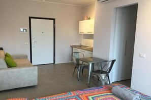 A modern living room with a sofa, a dining table with chairs, a kitchen area with cabinets, and a door.