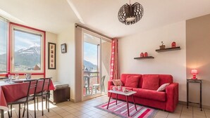 A living room with a red sofa, a dining area with a table and chairs, a balcony with a view of mountains, and a red lamp on a side table. - 150m from ski slopes, 4th floor, view mountain, balcony, tv, 31m², Serre Chevalier (Briançon)