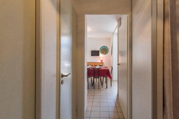 A narrow hallway leading to a dining area with a table and chairs, a television on the wall, and a light fixture.