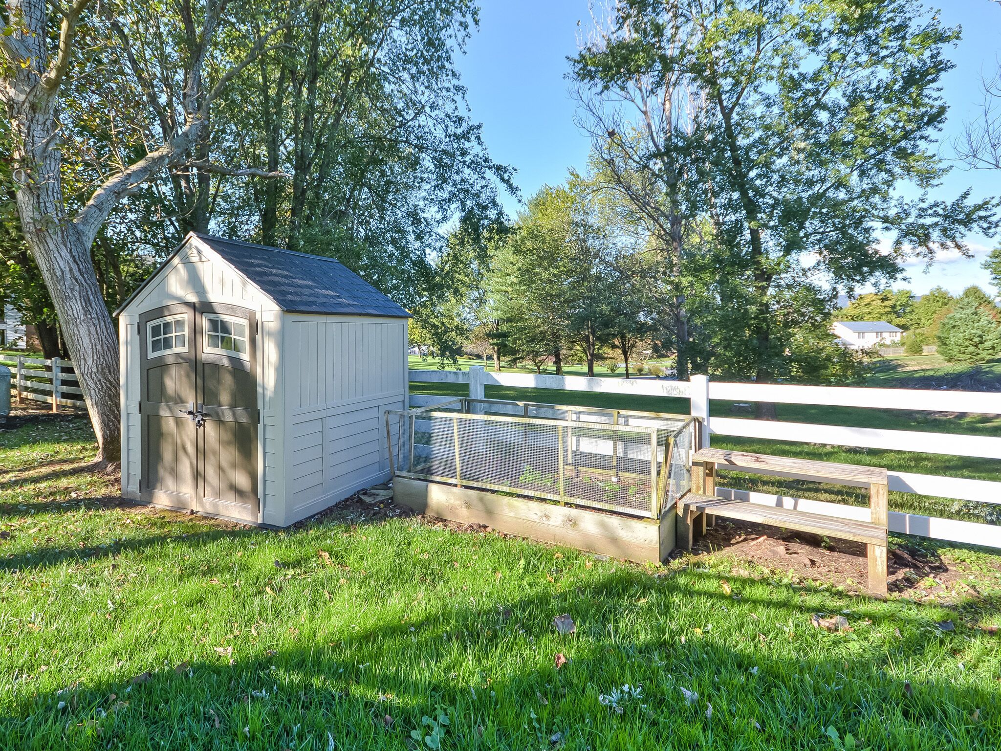 A small, white wooden shed with a door and windows, located next to a white fence in a grassy area with trees in the background.