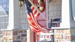 A porch with a stone wall, a red door, an American flag, a chair with a cushion, and a 'STAY' sign. - Charming "Cream of the Crop" Cape ~❤️of Middletown (Middletown)