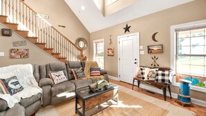 A spacious living room with a high ceiling, a staircase, a large window with a view, and a cozy fireplace area. - Charming "Cream of the Crop" Cape ~❤️of Middletown (Middletown)