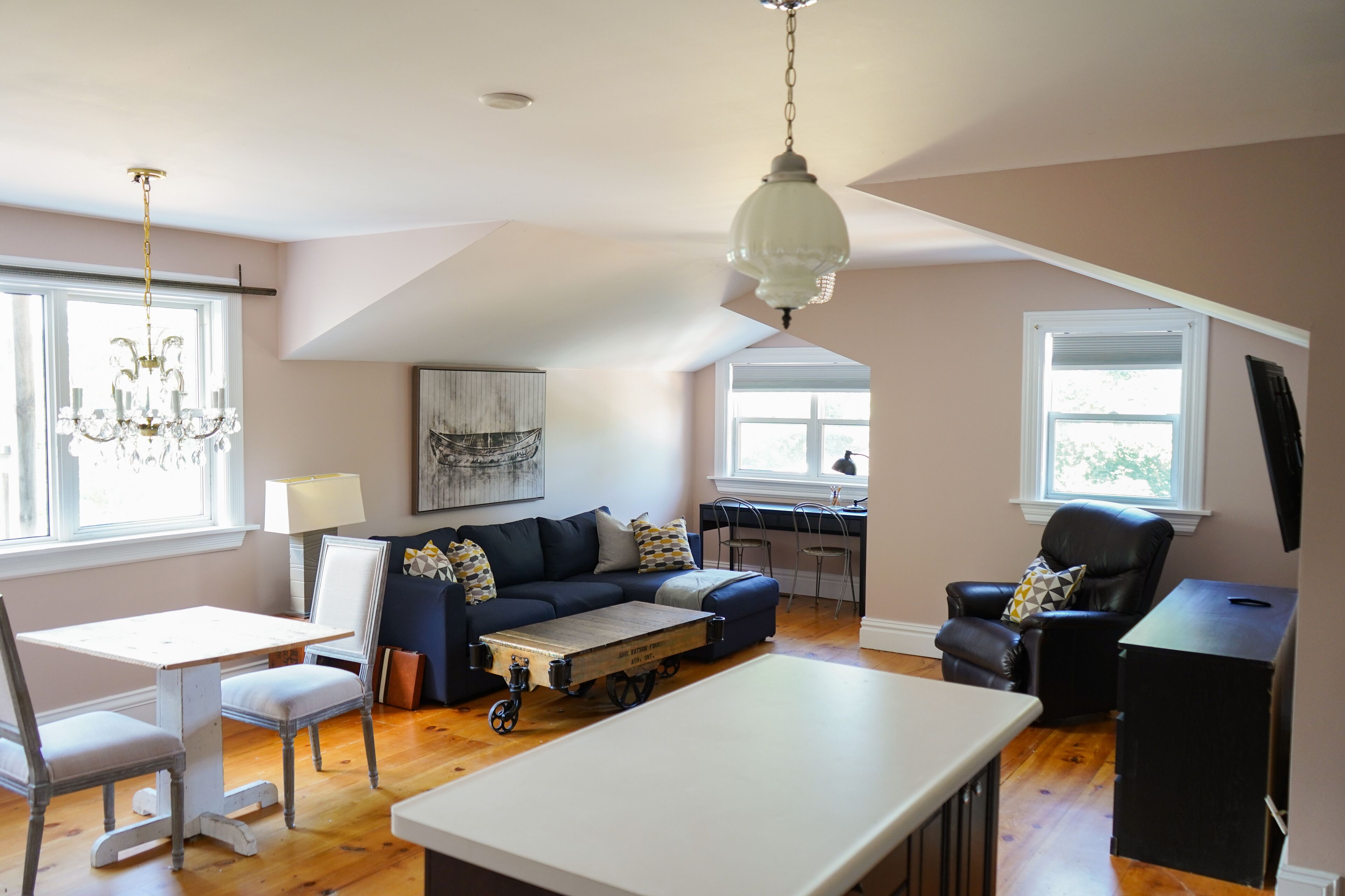 A spacious living area with a dark blue sofa, a white coffee table, and a dining table with chairs. There is a chandelier hanging from the ceiling and a window providing natural light.
