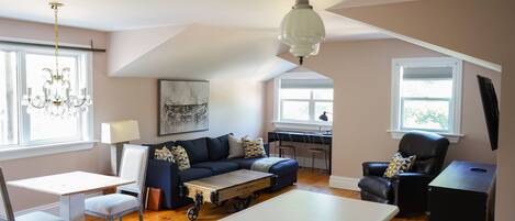 A spacious living area with a dark blue sofa, a white coffee table, and a dining table with chairs. There is a chandelier hanging from the ceiling and a window providing natural light.