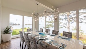 Un coin repas avec une table dressée pour six personnes, de grandes fenêtres donnant sur un lac et un lustre moderne.