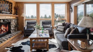 A cozy living room with a fireplace, two sofas, a coffee table, and large windows offering a view of the outdoors.