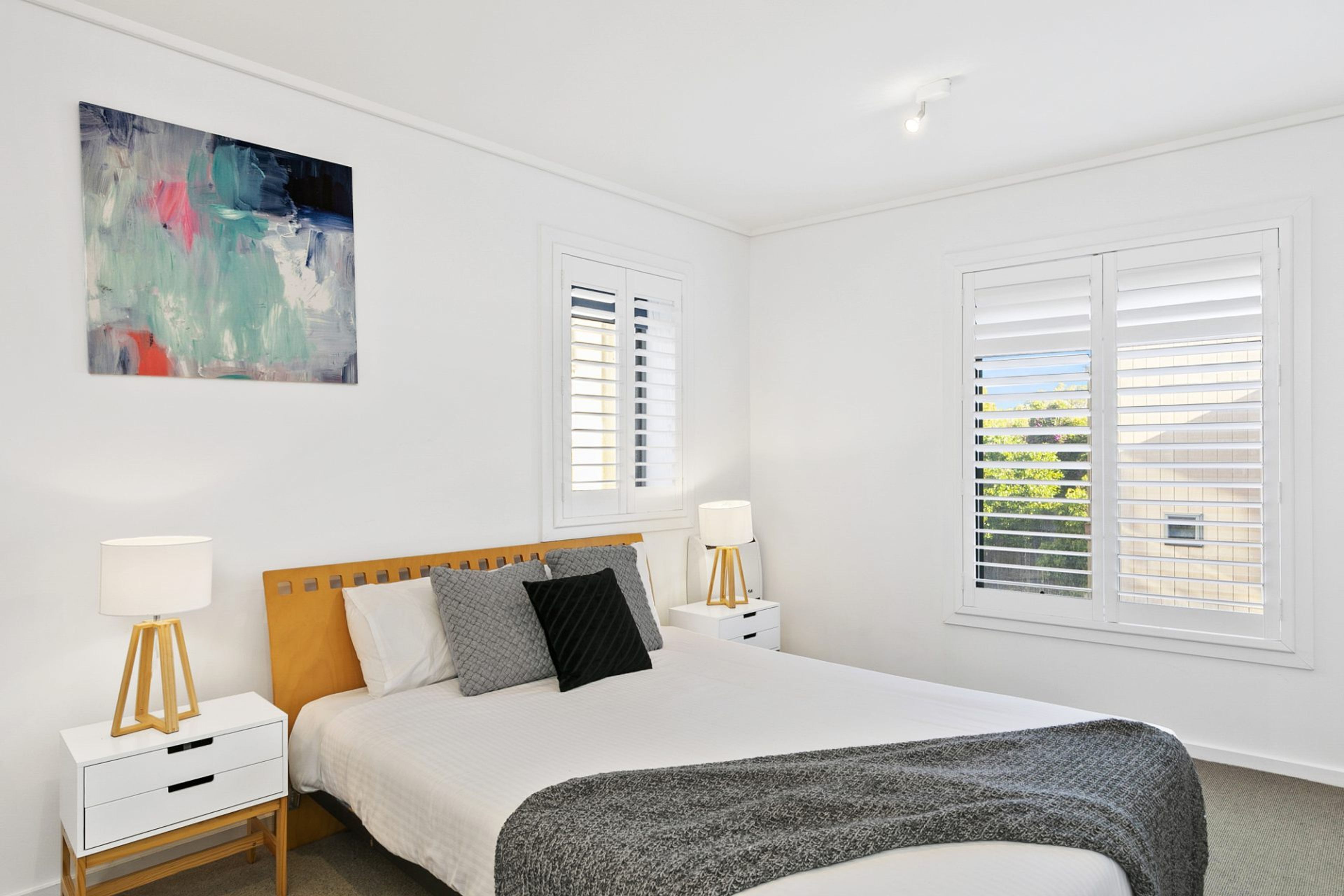 A bedroom with a bed, two bedside tables, a lamp, a painting, and a window with blinds.