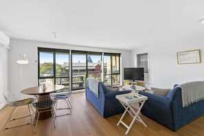 Un salon moderne comprenant un canapé, une table à manger, un téléviseur et de grandes fenêtres donnant sur des immeubles et des arbres.