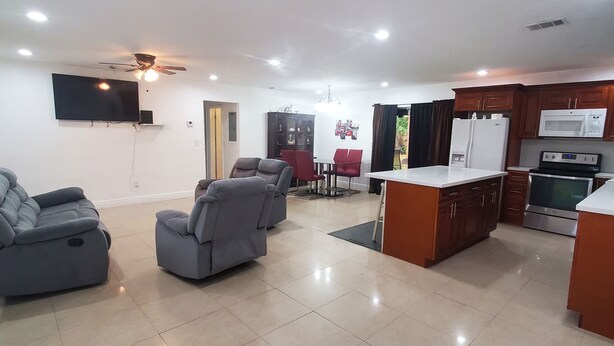 A spacious living area with a flat-screen TV, grey sofas, a ceiling fan, and a kitchen with modern appliances. - Jazzy Escapes (with pool) near Hard Rock Stadium (Miami)