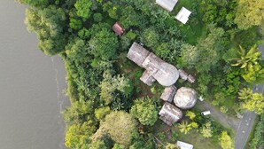Vista aérea de una propiedad con un amplio espacio abierto rodeado de abundante vegetación, incluyendo árboles y arbustos. Varias estructuras grandes y circulares con techos de paja, posiblemente utilizadas para reuniones o eventos. Un sendero pavimentado atraviesa la propiedad, y se observa una pequeña masa de agua a la izquierda.
