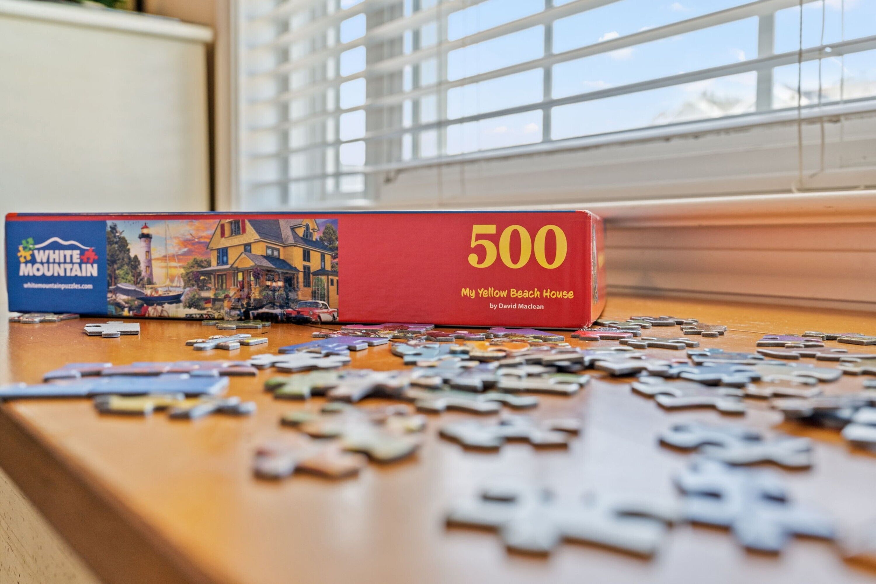 A puzzle box labeled "500 My Yellow Beach House" by David Maclean on a wooden table with scattered puzzle pieces.