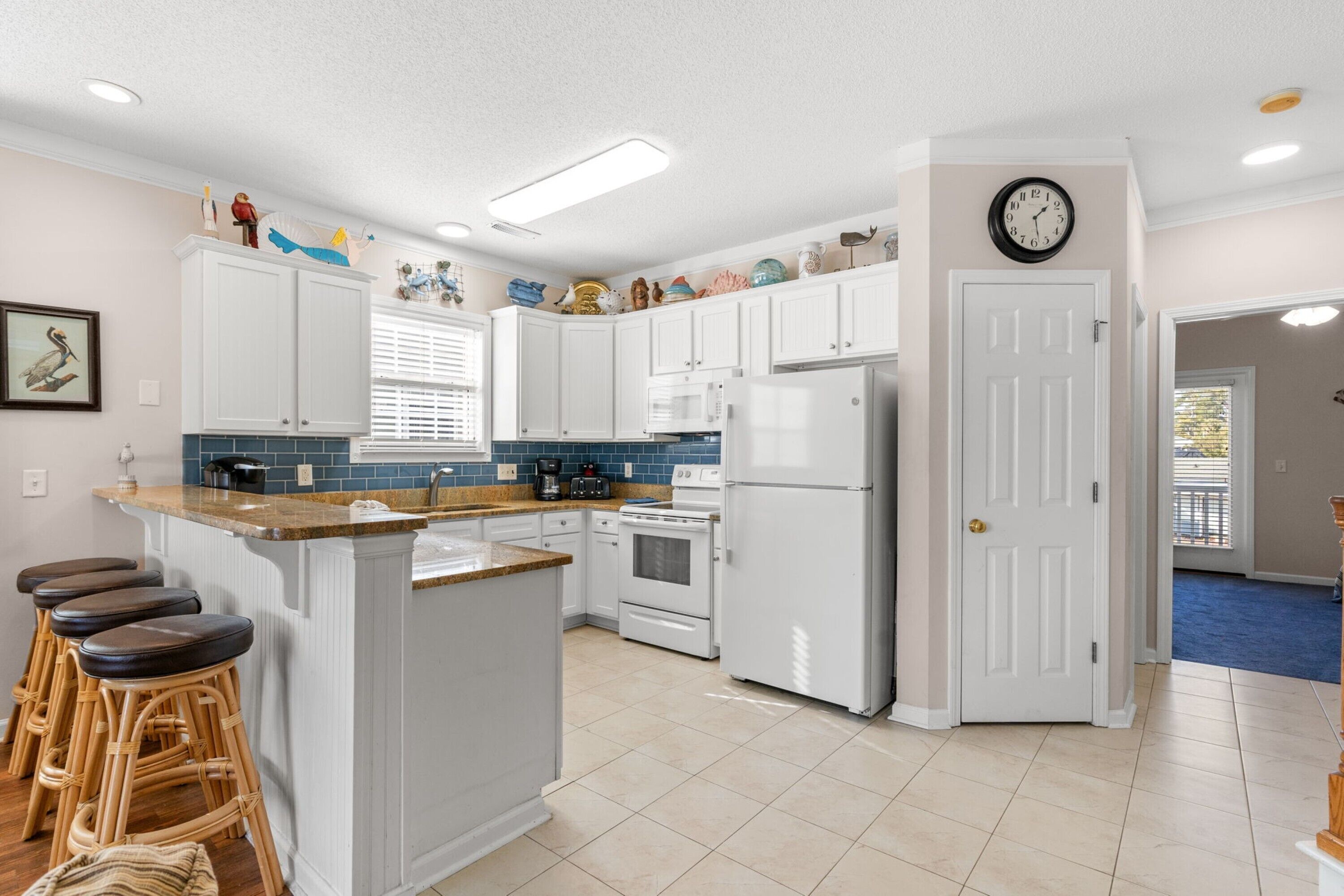 A kitchen with white cabinets, a central island with stools, a refrigerator, and a clock on the wall.