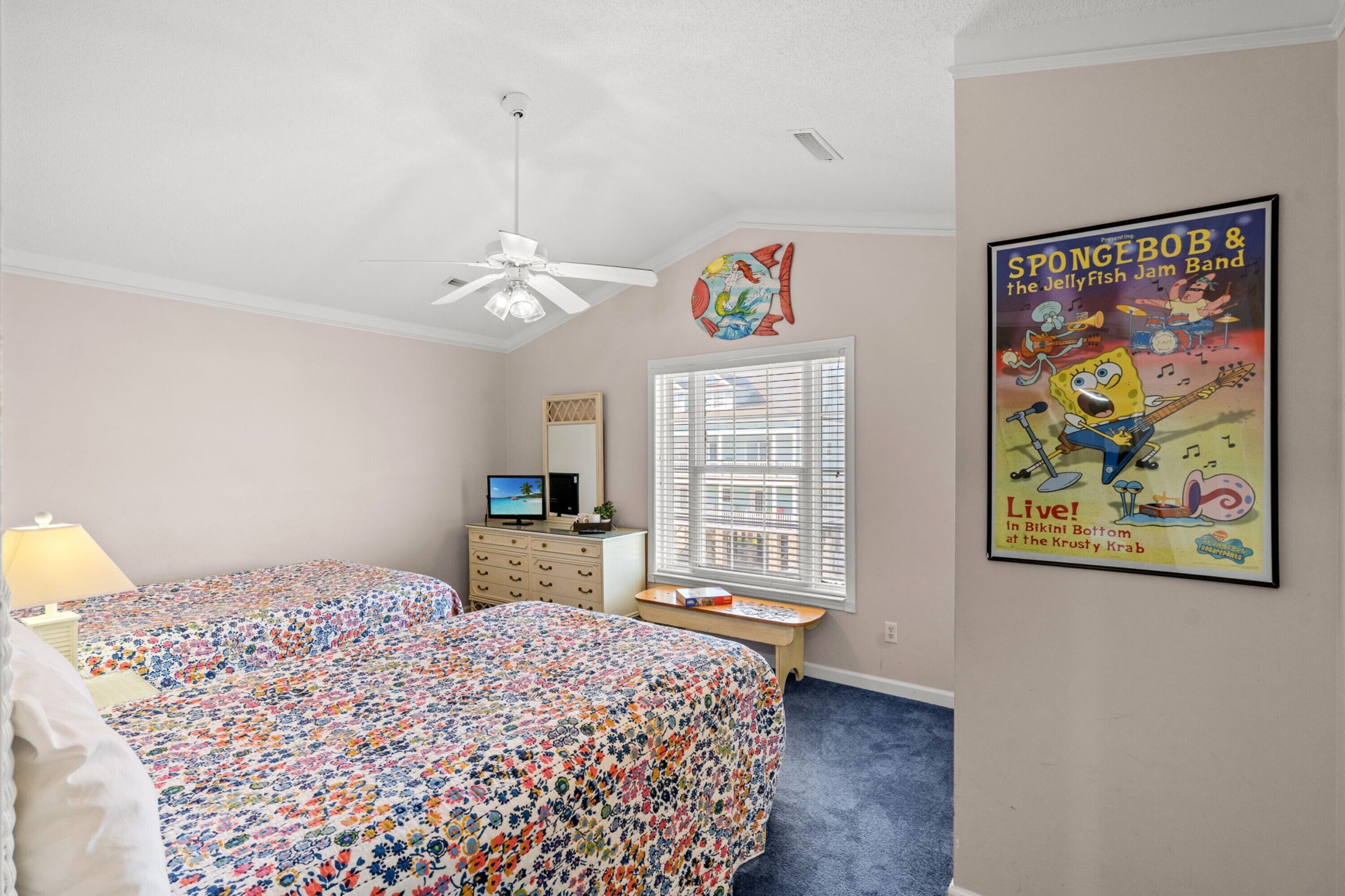 A bedroom with two beds, a ceiling fan, a dresser, a desk, a window with blinds, and a poster on the wall.