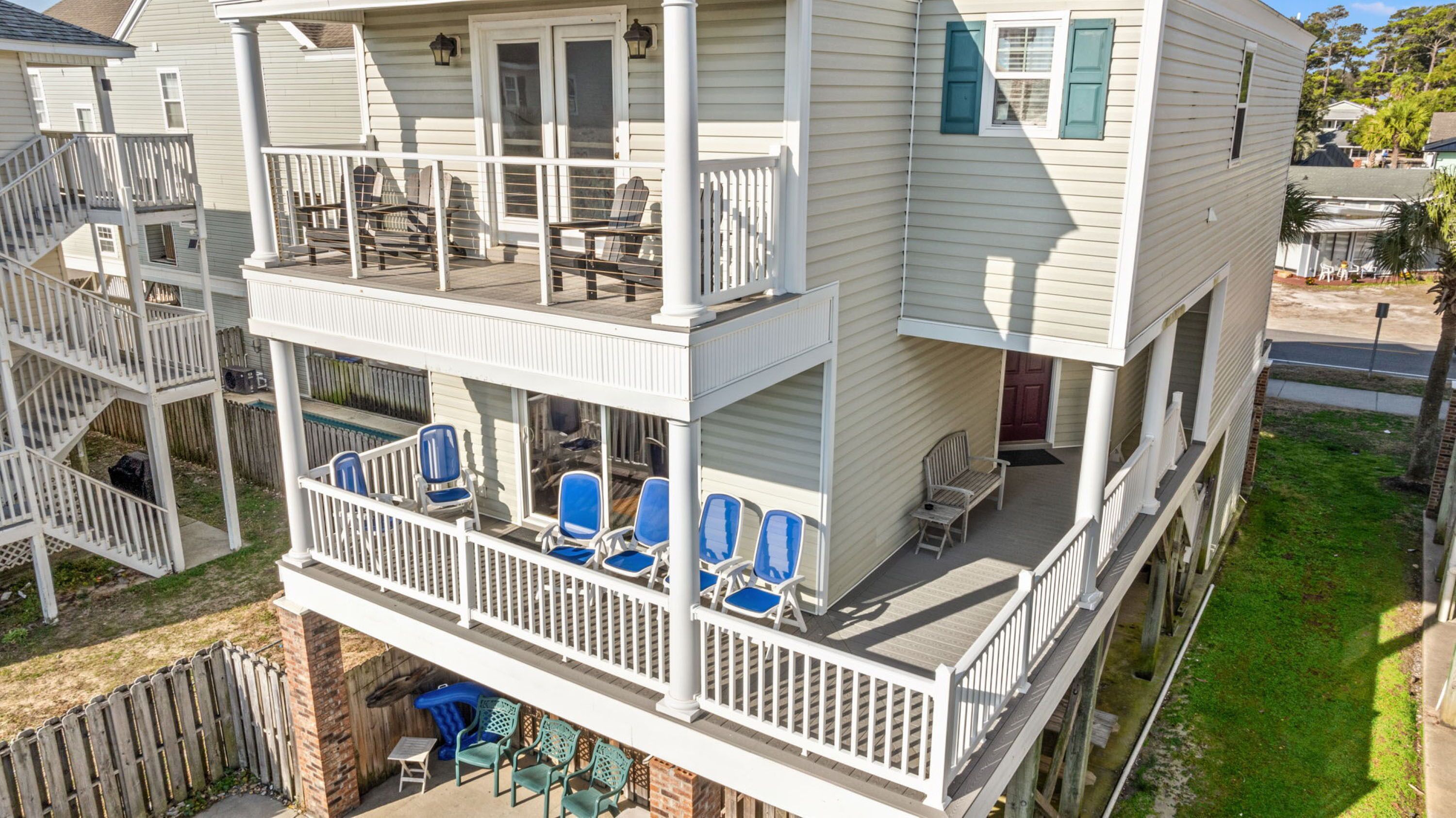 A multi-story building with a balcony on the ground floor, featuring several blue chairs and a small table.