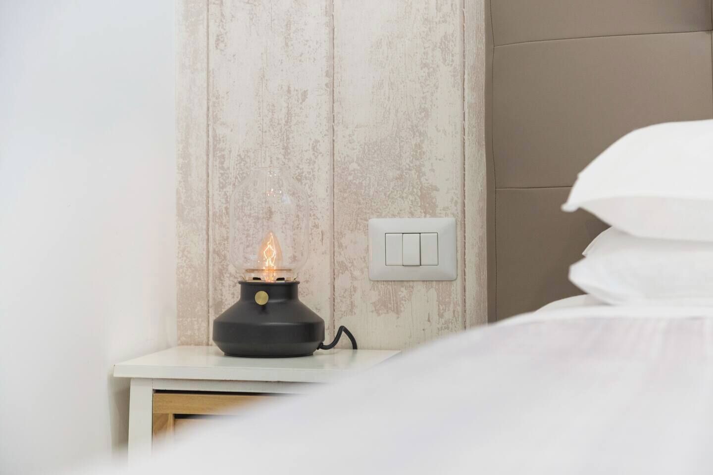 Une table de chevet avec une bougie allumée et un interrupteur, un lit avec du linge de lit blanc et un panneau mural en bois.