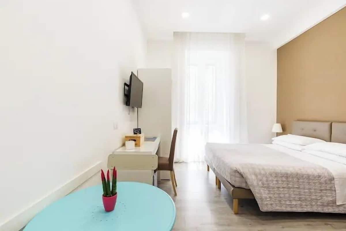 A hotel room with a bed, a desk with a chair, a TV mounted on the wall, and a potted plant.