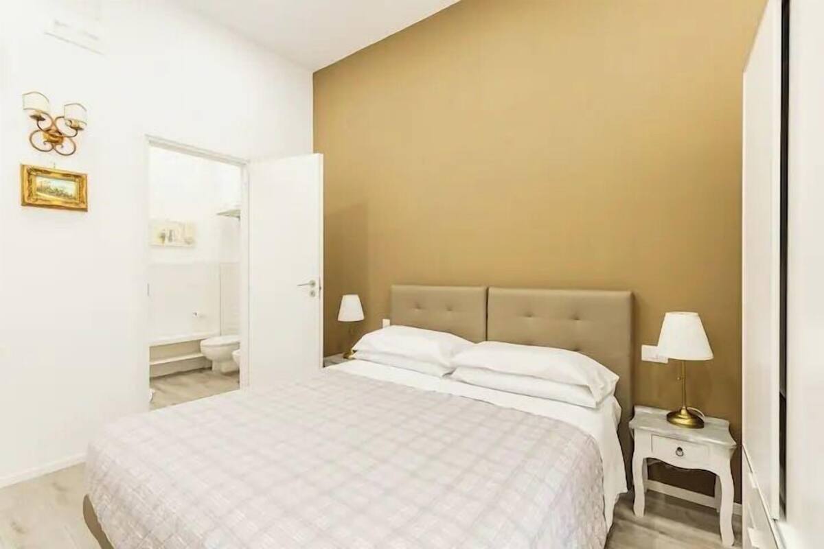 A bedroom with a bed, two bedside tables, a nightstand, a wall-mounted lamp, and a door leading to a bathroom.