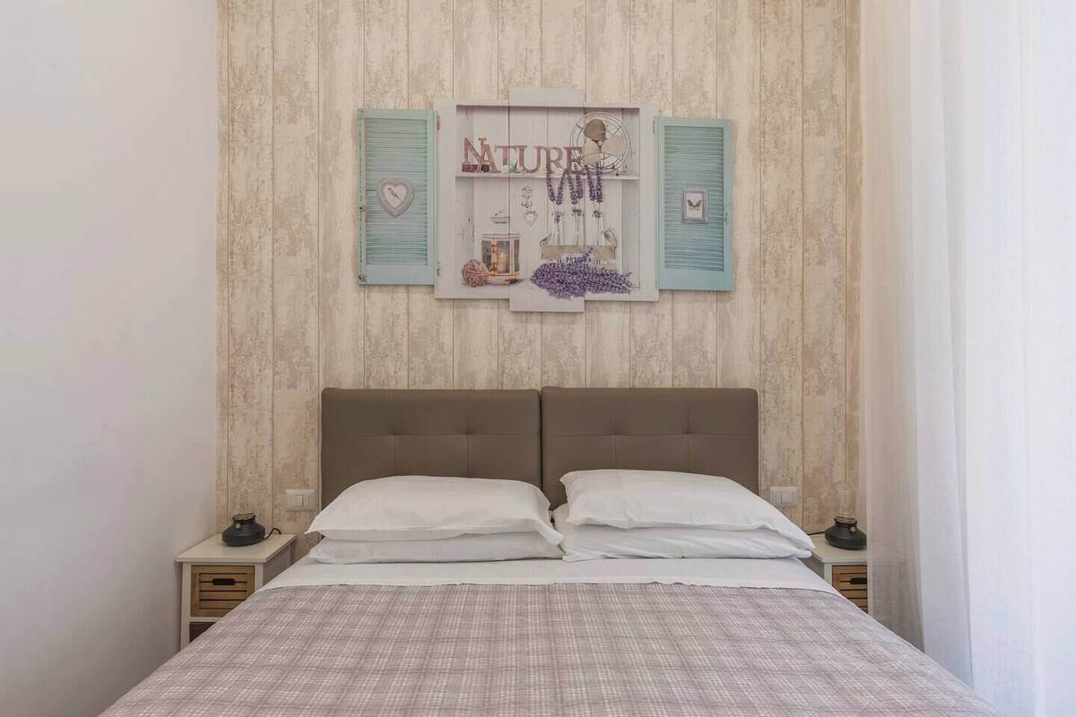 A bedroom with a double bed, two nightstands, a wall-mounted shelf with decorative items, and a wooden textured wall.