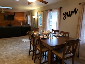 Un coin repas avec une table en bois, dressée pour quatre personnes, entourée de chaises. Un téléviseur est fixé au mur, et un ventilateur de plafond est installé au-dessus.