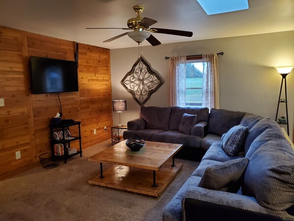 A cozy living room with a wooden wall, a ceiling fan, a flat-screen TV, a sofa, a coffee table, and a window with curtains. - Hilltop Retreat, a modernly-rustic getaway! (Murphysboro)